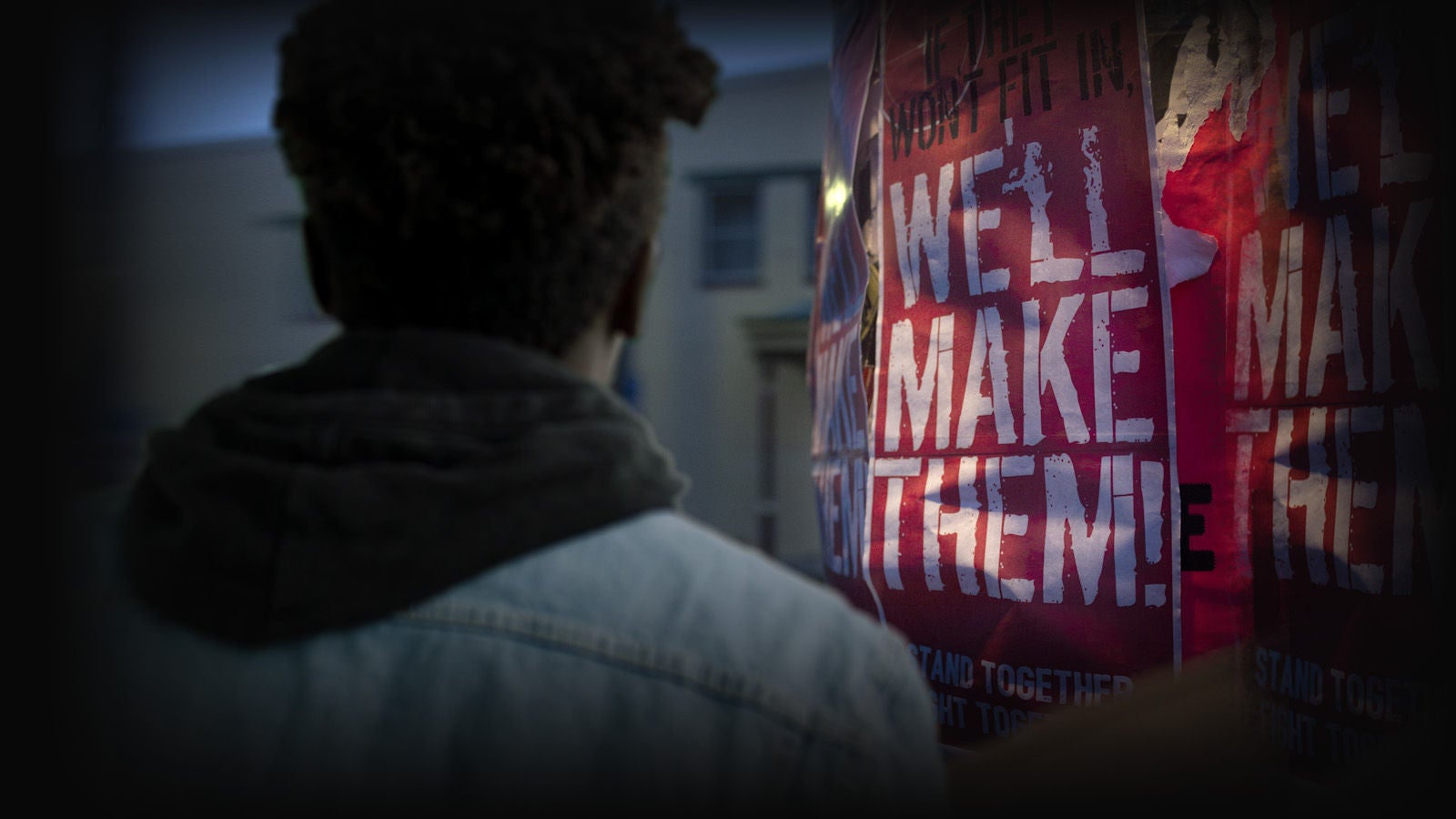 A man stands with back to camera. He is facing a pole that has been plastered with a poster that reads 'if they won't fit in, we'll make them'.